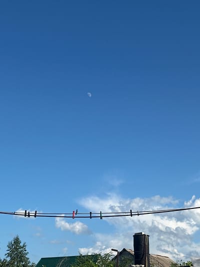 This is a vertically oriented, outdoor, daytime, medium shot photograph looking up towards the sky. The scene depicts a bright blue sky with scattered white clouds on the right side of the frame. A portion of the moon is visible as a small, pale crescent in the left half of the sky.
In the right portion of the image, a clothesline stretches diagonally upwards. Attached to the clothesline are several colorful clothespins: green, red, and dark colored ones. Behind the clothesline, a dark, cylindrical water tank is mounted on a structure, partially obscured by green foliage and the roof of a building. The building appears to have a corrugated metal roof.
The time of day appears to be daytime, likely late morning or early afternoon, given the bright sunlight and the visibility of the moon in the blue sky. The weather seems to be clear and pleasant, with only a few scattered clouds. The location context is Tobyl, Kazakhstan. No text is visible in the image.