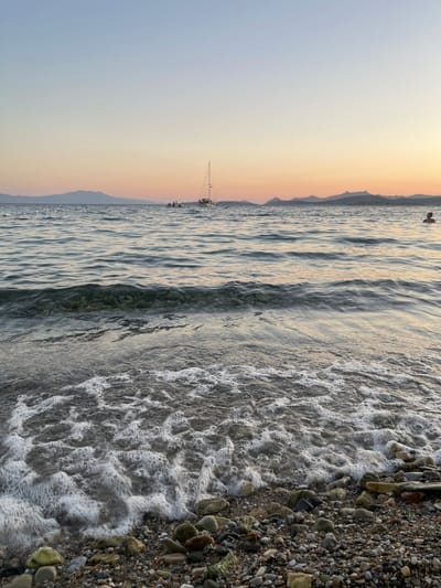 A low-angle, vertical, eye-level shot captures a tranquil beach scene at dusk. The foreground is dominated by a pebble-strewn shoreline, where a gentle wave with white foam recedes. The water, a gradient of blues and grays, exhibits soft ripples that reflect the soft orange and pink hues of the sunset sky. In the middle ground, a white sailboat with a tall mast is anchored, carrying a small group of people. Further in the distance, a chain of mountains creates a silhouette against the colorful horizon. A lone swimmer is visible in the upper right quadrant of the frame, with only their head and shoulders above the water. The overall atmosphere is serene and peaceful, indicative of a quiet evening by the sea in Bodrum, Turkey.