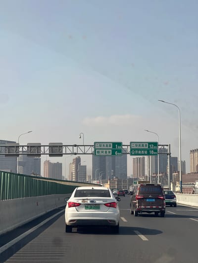 The image displays an elevated multi-lane highway in Jinan, Licheng District, China, under bright daylight. The scene is viewed from within a vehicle traveling forward.

In the immediate foreground, a white sedan with a green new-energy vehicle license plate "A·DF0988" occupies the middle lane, moving away from the viewer. To its right, in the adjacent lane, a dark reddish-brown van/MPV is present, displaying "FFQ188" as a reflection on its rear window and a license plate "鲁A FQ185". Multiple other vehicles, including cars and vans, are visible in all lanes ahead, receding into the distance.

Overhead, a metal gantry spans the highway, supporting two green rectangular road signs. The left sign displays "二环东高架" (Second Ring East Viaduct) at 1 km and "顺河高架" (Shunhe Viaduct) at 6 km. The right sign indicates "二环西高架" (Second Ring West Viaduct) at 12 km and "济南西站" (Jinan West Railway Station) at 18 km. Modern streetlights are mounted on poles along the highway.

To the left, a green vertical slat barrier borders the highway. The background reveals an urban skyline characterized by numerous high-rise buildings and skyscrapers, appearing under a light blue, slightly hazy sky.