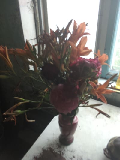 A bouquet of flowers, primarily orange lilies and deep red roses, is arranged in a vase. The vase, made of a reddish-brown material, sits on a white, speckled surface, possibly a windowsill or table. The background shows a blurred view through a window, with a hint of greenery and a light sky, suggesting it might be daytime. The lighting is soft, casting a gentle glow on the flowers. The overall impression is one of quiet beauty, with the flowers appearing full and vibrant despite the slightly blurry image quality. There is no visible text in the image. The scene appears to be indoors, near a window.