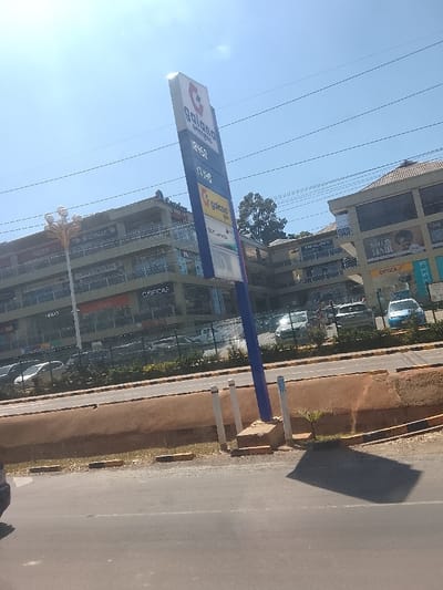 A tall sign for Galana Energies displays fuel prices (184.68, 171.10) and brand logos. Behind it, a multi-story building houses various businesses including Optica, Salon Hair, and Vivo. Cars are parked in front. A narrow strip of greenery separates the road from the building. A palm sapling grows near the sign's base. The image is captured on a bright day with blue skies.