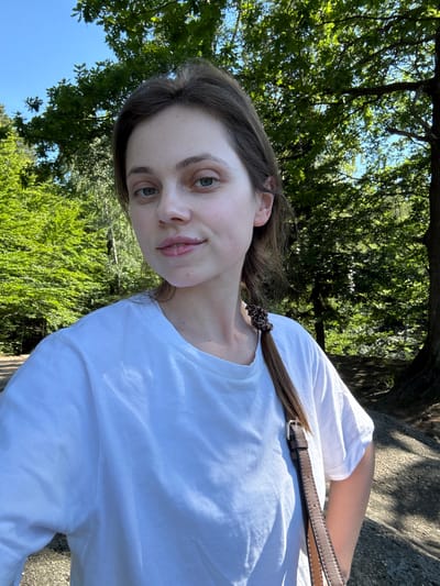 A young woman with brown hair tied in a braid and wearing a white t-shirt stands in a forest. She is looking at the camera and smiling slightly. The background shows trees and a bright blue sky. The woman has a brown bag strap over her shoulder. The location is the city of Szklarska Poręba, Poland.
