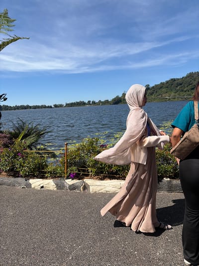 This is a full-shot, eye-level, outdoor photograph of two women walking on a paved path near a lake in Baturiti, Indonesia. The woman on the left is wearing a beige hijab and a long beige dress that is flowing in the wind. She is walking away from the camera, looking to her left. The woman on the right is partially visible from the waist down, wearing a teal shirt and black pants, and carrying a brown patterned crossbody bag. Her head and face are not visible. The background features a large body of water with some lily pads, a lush green hill covered in trees, and a bright blue sky with wispy white clouds. In the foreground, there is a decorative border made of grey and white stones. The overall scene depicts a serene and sunny day by the lake.
