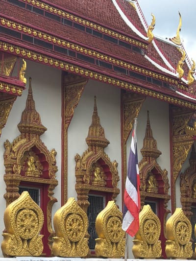 The image shows the exterior of a temple building, likely a Buddhist temple, in the city of Chang Phueak, Thailand. The architecture is ornate, featuring a red tiled roof with gilded decorative elements along the eaves. The walls are white, with elaborate gold and red accents above windows and along cornices. Several golden stupa-like structures with intricate carvings are visible, some containing small golden Buddha statues. In front of these, a series of golden decorative elements resembling lotus flowers with a Dharma wheel symbol are mounted on a railing. A Thai flag, with its red, white, and blue stripes, is prominently displayed on a flagpole in front of the building. The sky above is overcast. The overall impression is one of religious architecture characterized by rich ornamentation and symbolism.