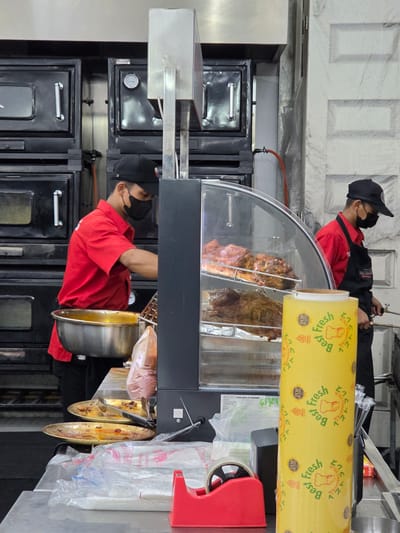 This image captures a scene within a food establishment, likely a restaurant or food stall in Medan, Indonesia. The primary subjects are two individuals, both wearing face masks and red uniforms, suggesting they are employees. One individual, in the foreground, is wearing a black cap and a red shirt. They are leaning over a large, cylindrical metal pot, possibly preparing or serving food. A second individual, in the background and also wearing a black cap and red shirt, appears to be interacting with a large roll of yellow plastic wrap.
The setting is a commercial kitchen or food preparation area. Visible objects include kitchen appliances such as ovens or warming units, a glass display case showcasing large cuts of meat, and various containers and utensils. The yellow plastic wrap prominently features text in both English and what appears to be Japanese characters, along with graphics indicating "Best Fresh," "TOP BRAND," and "HALAL." This suggests the establishment caters to a diverse clientele and emphasizes the freshness and religious compliance of their products.
The overall impression is one of a busy food service environment. The lighting suggests an indoor setting, likely during operating hours. The presence of the meat in the display case and the preparation activities indicate that food is actively being handled and served. There are no strong emotional cues visible due to the face masks, but the actions suggest a focus on work and customer service. The visible text on the packaging provides brand and product information, reinforcing the commercial nature of the scene.