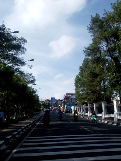 The image depicts a street scene in Phou Khoun, Laos. The perspective is from a pedestrian crosswalk. There are several motorbikes travelling down the road. The road is bordered by trees on both sides, with a fence on the right. The sky is blue with some clouds. Street lamps are visible along the road. Buildings and billboards are visible in the distance. People are walking on the sidewalk to the left of the street.