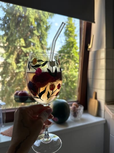 A person holds a glass with a clear straw, filled with liquid and what appears to be fruit, possibly strawberries, against a window backdrop. The glass is decorated with floral designs. Outside the window, there is green foliage and glimpses of a bright sky. The scene appears to be indoors, near a window, possibly in a kitchen or dining area. In the background, kitchen items such as a cutting board, fruit, and a kitchen counter are visible. There is no clear indication of the location being Kiel, Germany, from the image alone.