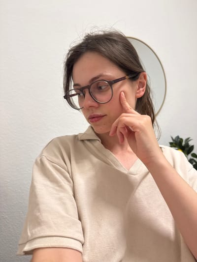 A woman with brown hair and wearing glasses is touching her cheek with her right hand. She is wearing a light beige long-sleeved shirt. Behind her is a white wall, a round mirror, and a plant. It is not possible to determine the location as Kőröshegy, Hungary, based on the visual information provided.