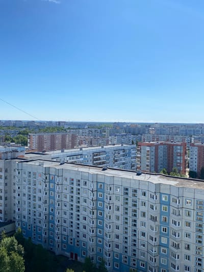This is a vertical, eye-level, full shot of a sprawling cityscape under a clear blue sky. The perspective is from a high vantage point, looking out over numerous Soviet-era apartment buildings stretching towards the horizon. The buildings are predominantly grey and blue, with many windows visible. Some buildings have reddish-brown brick facades. The scene depicts an urban environment with a dense concentration of high-rise residential blocks, interspersed with some greenery. The time of day appears to be midday or early afternoon, given the bright sunlight and clear sky. The weather seems to be fair and sunny. There are no people or distinct objects visible in the foreground, nor is there any text. The overall impression is one of a vast, somewhat monotonous urban landscape typical of many cities in Russia. The lack of activity or any discernible event suggests a passive observation of the city's layout. The overall mood conveyed is one of scale and the uniformity of urban living.