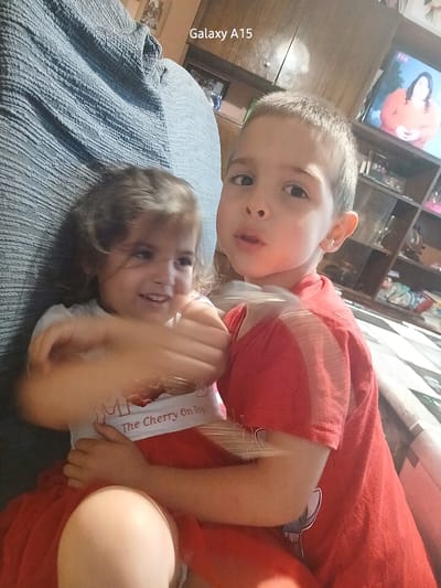 The image features two young children, a boy and a girl, engaged in a playful interaction on a bed or sofa in an indoor setting. The boy, wearing a red t-shirt, is in the foreground, lying on top of the girl and looking directly at the camera with a neutral expression. The girl, positioned beneath him and also wearing a red outfit with a white undershirt, smiles broadly towards the side with her arms raised, conveying a sense of joy or playfulness.
They are resting on a textured blue blanket or sheet. In the background, a brown wooden furniture piece, possibly a shelf, is visible. A television screen can be seen displaying what appears to be an orange cartoon character, possibly a fox.
Notable details include the text "Galaxy A15" written vertically along the left edge of the image, indicating the camera device. The girl's white undershirt prominently displays the phrase "The Cherry On Top." The lighting suggests an indoor scene, likely during the day or under artificial light. The overall mood is cheerful and relaxed. The location context for this scene is Obrochishte, Bulgaria.
