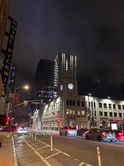The image depicts a street scene at night in San Francisco, California, United States of America. Dominating the foreground are several vehicles, including a silver SUV, a dark sedan, and a gray hatchback, stopped at a traffic light. The street is wet, reflecting the city lights and traffic signals, suggesting recent rain.
In the background, an iconic white building with arched windows and a prominent clock tower stands illuminated. Above it, a modern, dark glass skyscraper rises into the hazy night sky, its upper floors lit with vertical bands of light. To the left, a tall, vertical sign reads "HOTEL PICKWICK." Traffic lights show red, indicating that vehicles are at a standstill. The overall atmosphere is urban and dynamic, with a mixture of historic and contemporary architecture.
