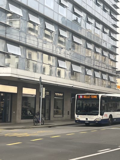 An urban street scene in Geneva, Switzerland, features a white and blue public transport bus, a modern glass-fronted building, and a paved road with markings. The image appears to be taken during the daytime under clear weather conditions.
The prominent subject is a Mercedes-Benz public bus, operated by "TPG" (Transports Publics Genevois), displaying "9 BELLE-TERRE" on its front destination sign, indicating Line 9 heading towards Belle-Terre. The bus is stationary on the right side of the frame, facing slightly left.
To the left of the bus is a multi-story, contemporary building characterized by its reflective glass facade, which mirrors the classical architecture of other buildings across the street. Many of the small rectangular windows on the building's exterior are slightly open or tilted outwards. On the ground floor of this building are several shopfronts; one clearly displays "PASTORE-NICOLET" and another appears to read "LA FRIE STUDIO".
The foreground shows a paved street with distinct yellow dashed lines and a white solid line. A traffic light pole, to which a black bicycle is securely locked, stands between the bus and the building. The overall impression is a bustling city environment, typical of a European urban center, with a mix of modern and traditional elements reflected in the architecture.