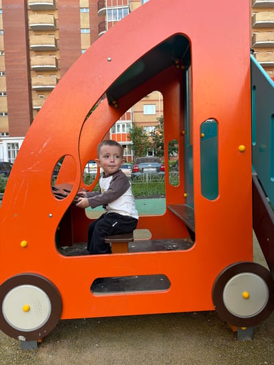 A young Caucasian boy with short brown hair and light skin is sitting inside a bright orange children's playground structure designed to resemble a vehicle. He is wearing a long-sleeved shirt with a white body and brown sleeves, and dark corduroy pants. The boy is holding onto a steering wheel-like element, looking to his right with a slightly open mouth. The playground structure features yellow spherical decorative elements and a gray seat. In the background, there is a tall, multi-story apartment building with brick and yellow facade, and balconies. Several cars are parked near the building. The ground is covered with small pebbles and sand. The setting appears to be a residential courtyard in the city of Vniissok, Russia.