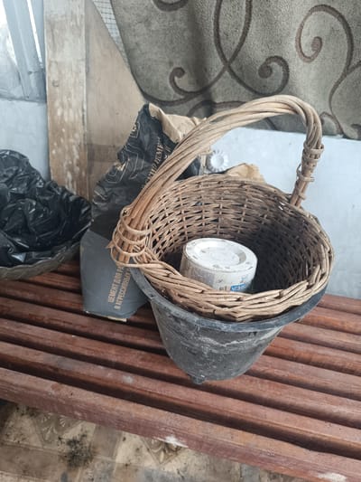 A close-up, eye-level, indoor shot focuses on a woven basket with a black plastic bucket liner, resting on a wooden bench. Inside the basket is a white cylindrical object, possibly a can or container, with some visible Russian text and a percentage symbol. To the left of the basket, a black garbage bag is partially visible in a wicker basket. Behind the woven basket, a large, dark bag with white Russian text, appearing to be a bag of cement, is propped up. The background features a textured fabric with a swirling pattern and a piece of weathered wood. The overall scene suggests an outdoor or utilitarian setting, possibly a shed or covered porch, with evidence of dust and dirt on the bench and objects. The image is taken in Ostrogorka, Russia.