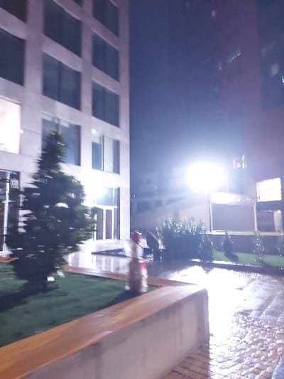 This nocturnal image captures an outdoor urban environment, likely a courtyard or pedestrian area, in Tiranë, Albania. The photograph is oriented vertically (originally taken in landscape and rotated left), with a dark night sky dominating the upper left quadrant. A prominent, bright artificial light source, possibly a streetlamp, casts a significant glare in the upper center, illuminating parts of the scene and reflecting on wet surfaces.
On the right side of the image, a modern building with a facade of evenly spaced rectangular windows stretches from the bottom towards the center. In the foreground, a long, light-colored planter box or seating structure with a wooden side extends horizontally, partially framing the scene. Adjacent to this structure is a paved area that is visibly wet and reflective, indicating recent rainfall.
Near the planter, positioned on the wet ground, is a blurry, upright object with distinct red and white stripes or sections, topped with a dark rim. Its exact function is unclear, but it appears to be a decorative or functional element of the public space, such as a bollard or a small planter. Patches of green landscaping, resembling artificial turf or well-maintained grass, are interspersed within the area. A small tree or bush with dark foliage is also visible towards the lower middle of the image, partially obscured by blur. No discernible human figures or specific activities are present, and there is no visible text in the frame. The overall impression is one of a quiet, illuminated urban space after rain.