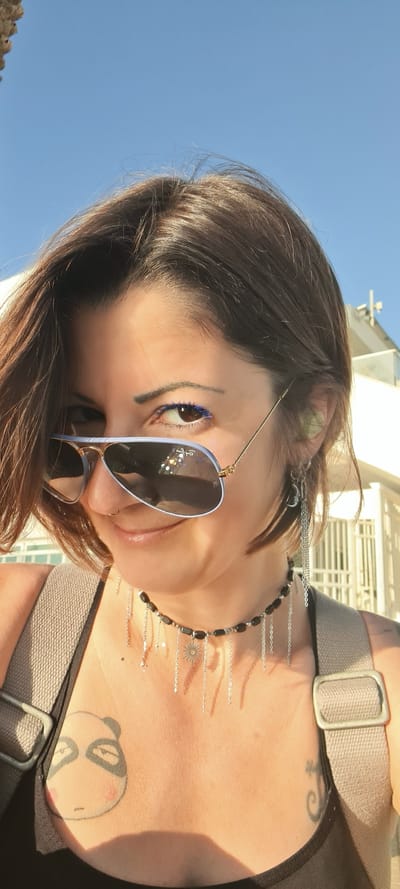 A woman wearing sunglasses and a black necklace is looking at the camera. The background features a bright blue sky and the top of a building. She is wearing blue mascara. The picture was taken in Nahariyya, Israel. The woman has several tattoos including one of a cartoon animal.