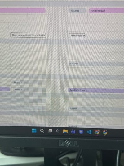 The image displays a close-up, slightly angled view of a computer monitor, focusing on its screen content and the bottom bezel. The screen presents a grid-based interface, resembling a spreadsheet or calendar application, with horizontal and vertical lines defining cells. Various cells contain text, primarily "Absence" in gray boxes. Other visible text includes "Absence (en attente d'approbation)", "Absence (en at", "Revolte Noyal" (in a purple highlighted cell), and "Revolte St Priest" (in another purple highlighted cell). Below the application interface, a Windows 11 taskbar is visible, displaying icons for the Start menu, Search, File Explorer, Microsoft Teams, Discord, Visual Studio Code, and Google Chrome. The physical bottom bezel of the monitor is black and features a prominent "DELL" logo centered below the screen. The monitor's surface reflects ambient light, and indistinct darker shapes are visible beneath the monitor, suggesting a desk environment. No individuals are present. The precise location within the city of Thiais, France, cannot be determined from the visual information provided in the image.