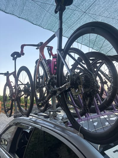 The image depicts a silver car parked on a paved surface, possibly a driveway or parking lot, with a multi-bike rack mounted on its roof. Three bicycles are loaded onto the rack, with their wheels facing outwards. The bicycles appear to be road bikes, featuring thin tires and drop handlebars. One bicycle, in the foreground, is a dark color with red accents on the handlebars and frame. Behind it, another bicycle is visible, predominantly red. A third bicycle, partially obscured, has a frame with orange and pink hues and a pink water bottle attached. The scene is illuminated by natural light, suggesting it is daytime. A green mesh shade covers part of the upper frame, and the sky is visible in the background, indicating an outdoor setting. The overall impression is that of a vehicle prepared for a cycling excursion. The location is specified as Dolenje Grčevje, Slovenia.