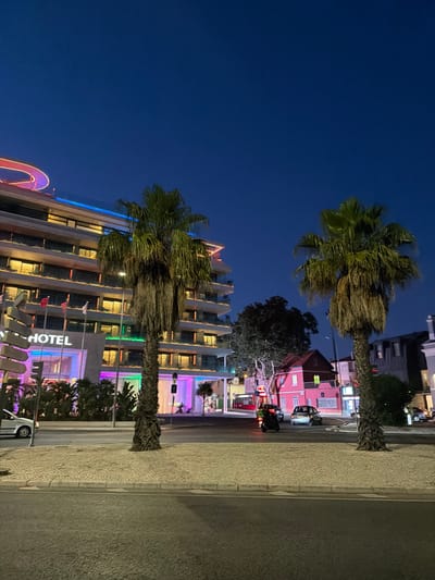 This media file depicts a nighttime scene in Cascais, Portugal. The image features a modern hotel building with illuminated windows and colorful exterior lighting, including blue and purple hues. Palm trees are prominent in the foreground, their fronds swaying, suggesting a breeze. A sidewalk with a textured surface runs alongside a road where a few vehicles, including a motorcycle and a car, are visible. In the background, other buildings, including a pink one, are also lit up. The sky is a deep blue, indicating it is nighttime. The overall atmosphere suggests a lively, urban setting, possibly a resort town or a vibrant city street. There is no clear indication of people engaging in specific activities, but the presence of vehicles and illuminated buildings points to ongoing activity. Visible text includes "_HOTEL" on the hotel building. The time of day is clearly night, and the weather appears clear.