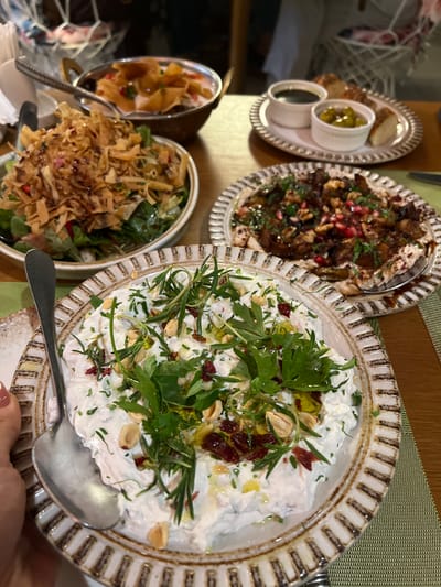 A close-up overhead shot captures a spread of Middle Eastern cuisine on a wooden table, likely in Abu Dhabi, UAE. In the foreground, a creamy white dip, possibly labneh or yogurt-based, is served in a decorative ceramic bowl. It's garnished with fresh green herbs, chopped nuts, and what appear to be dried cranberries or pomegranate seeds. A silver spoon rests partially in the dip. Behind this, to the left, is a salad with mixed greens topped with crispy fried pita bread and a drizzle of dark sauce. To the right, another dish features roasted vegetables, possibly eggplant, with a topping of walnuts and pomegranate seeds, served on a similar decorative plate. Further back, a small bowl contains a lighter colored dip with pieces of fried pita bread submerged in it. Next to that, a plate holds small white bowls of what seem to be a dark sauce, olives, and bread slices. The scene is set on a textured green placemat, with the background blurred, hinting at a restaurant setting.