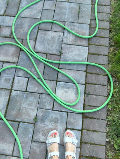 A high-angle shot captures a person's feet wearing white sandals with buckles, standing on a paved patio. The sandals have bright orange nail polish on the toes. A bright green garden hose is coiled haphazardly across the patio, with a section of lush green grass visible along the right edge of the frame. The patio is made of rectangular gray and beige pavers, with some moss and small weeds growing in the cracks. The lighting suggests it is daytime, likely outdoors. The scene appears to be a residential backyard or garden area in Fanipal, Belarus. There is no visible text in the image.