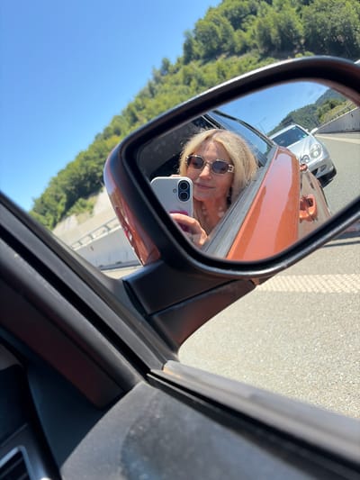 A woman with blonde hair and sunglasses takes a selfie in the reflection of a car's side mirror while driving on a sunny day. She is holding a white smartphone with a dual camera. The car appears to be copper-colored. In the background, through the car window and reflected in the mirror, a silver car is visible on a highway with trees and a concrete barrier. The setting is likely a highway outside of a city, given the road and surrounding landscape.