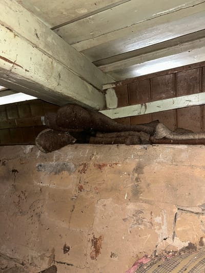 The image depicts an indoor scene, likely within an attic or crawl space, given the visible wooden beams, paneling, and concrete wall. The primary subjects are what appear to be multiple wasp nests hanging from a pipe or conduit. These nests are large, bulbous, and dark brown, with a textured, fibrous outer layer. They are clustered together, with one nest appearing to be composed of two fused sections.
The setting is rustic and appears somewhat neglected, with areas of rough concrete and chipped paint on the wooden structures. The lighting is dim, suggesting it's either nighttime or the space is poorly lit. There are no people visible in the image, and therefore no activity involving humans is taking place. The focus is entirely on the natural structures of the wasp nests and their immediate environment.
Notable details include the texture of the nests, the way they are suspended, and the rough condition of the surrounding materials. The context provided places this scene in the Відзаўскі сельскі Савет, Belarus, which gives a geographical location but does not offer specific visual cues to confirm it within the image itself. There is no visible text in the image. The overall impression is one of a hidden, potentially undisturbed natural formation within a man-made structure.