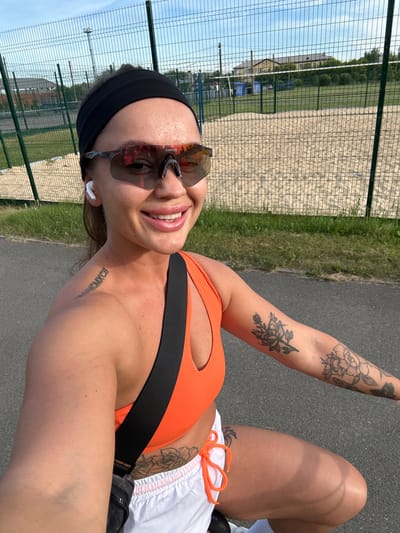 In Yuzhnouralsk, Russia, a woman is captured in a selfie outdoors during the day. She is wearing a black headband, sports sunglasses with reflective lenses, and white wireless earbuds. She has a black strap from a bag or accessory across her chest, and is wearing an orange sports bra and white shorts with orange drawstrings. Tattoos are visible on her left shoulder blade (Roman numerals) and her right arm (floral designs) and upper thigh. The background includes a chain-link fence, a sandy court area that appears to be a volleyball court, green grass, and some buildings in the distance under a blue sky. The woman is smiling and looking directly at the camera.
