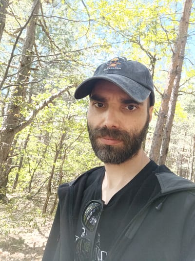 A middle-aged man with a beard is standing in a forest. He is wearing a dark-colored baseball cap with a logo, a black t-shirt, and a black jacket with sunglasses hanging from it. The forest has tall, thin trees with green leaves. The location is Sini Vrah, Bulgaria.