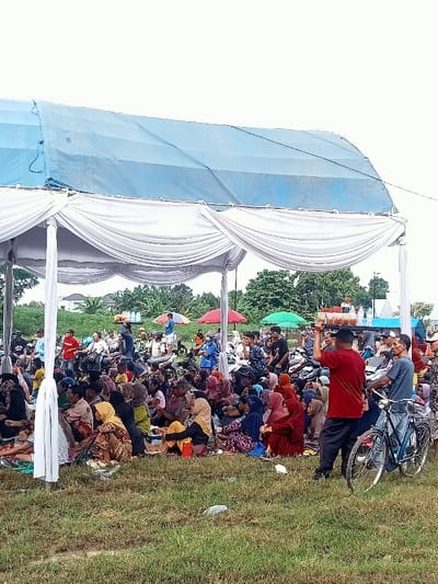 The image depicts a large gathering of people under a white canopy tent with a blue roof in a grassy field. The tent is supported by white poles and features white curtains along the sides. Numerous people are seated on the ground under the tent, many wearing traditional clothing. They are surrounded by a variety of objects including motorcycles, bicycles, and various personal belongings. A few individuals are standing and interacting with each other, and one person is balancing a tray of beverages on their head. In the background, there are several colorful umbrellas and a few buildings. The scene is located in the city of Kecamatan Margorejo, Indonesia, at latitude -6.736 and longitude 111.016. The environment is a field with green grass and a cloudy sky.