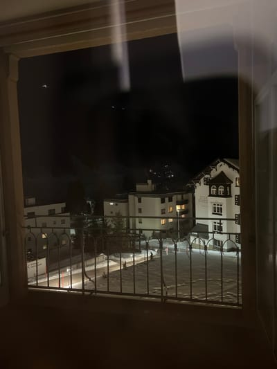 A view through a window, likely from an apartment or hotel room, overlooks a snowy street at night. Buildings with illuminated windows line the street, partially obscured by metal railings. The scene is illuminated by streetlights, casting a soft glow on the snow-covered ground and buildings. Reflections appear on the window's surface.