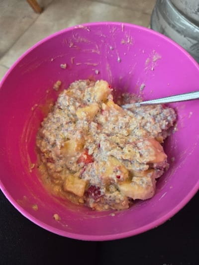 A magenta plastic bowl containing a creamy food mixture is depicted from a high-angle perspective. The mixture consists of a base resembling oatmeal or porridge, densely interspersed with small, dark, possibly chia seeds, and irregularly cut chunks of yellow fruit, consistent with banana or apple. Smaller fragments of red fruit are also visible throughout the mixture. A silver-colored spoon is partially submerged within the food on the right side of the bowl. Streaks and residue of the food adhere to the inner walls of the bowl, suggesting prior consumption or stirring. The immediate foreground beneath the bowl is an indistinct dark surface. The blurred background reveals a light brown object, possibly a wooden furniture leg, in the upper left, and a metallic, cylindrical object with a textured surface in the upper right. The scene is situated in the city of Gevgelija, North Macedonia.