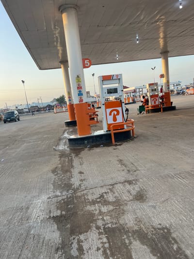 The image depicts a street view of a gas station in Bauchi, Nigeria. The scene is dominated by the open-air canopy of the gas station, supported by large, white pillars. Several gas pumps are visible, with an orange and white color scheme. One pump in the foreground has a digital display showing "LITRES," "SALE," and "PRICE." Another pump to the right has an orange fire extinguisher attached. In the background, other gas stations, buildings, and a street with traffic are visible. A dark colored car is parked on the left side of the road. Several people are present at the gas station, some appearing to be attendants or customers. One person is sitting on an orange bench near a gas pump, looking at their phone. The ground is paved with concrete, showing signs of wear and some wet patches. The sky is clear with a warm, hazy light, suggesting it is either early morning or late afternoon.