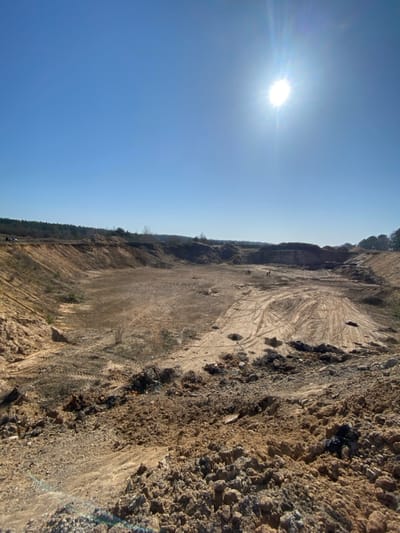 The image depicts a large, open-pit excavation or quarry site under a clear, bright blue sky in Верцялішкаўскі сельскі Савет, Belarus. The sun, a prominent bright flare with visible rays, is positioned in the upper right quadrant of the sky. The terrain consists of extensive disturbed earth, predominantly in shades of tan and brown, forming steep slopes and a relatively flat, expansive pit floor. Piles of excavated material are visible along the edges and within the pit. Numerous tire tracks crisscross the pit floor, particularly in the middle-right, indicating recent heavy machinery activity. Sparse, dry vegetation, including small bushes, is present on some slopes and parts of the pit floor. In the middle ground of the pit floor, two small, light-colored objects, possibly pieces of construction machinery, are faintly discernible. A dense line of trees, appearing as a forest, borders the site on the distant horizon, visible above the upper edges of the quarry. No human figures are clearly visible.