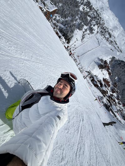 The image presents a first-person, tilted view of an individual on a snow-covered ski slope within the Les Allues commune in France. The central subject, an adult male, is positioned in the foreground, captured in a selfie. He wears a white quilted ski jacket over grey and black layers, bright lime green ski trousers, and a black and white 'Atomic' branded ski helmet with goggles resting on top. A snowboard is partially visible to his left. He has his eyes closed and a slight smile, with his right arm extended to hold the camera. The expansive, well-groomed slope exhibits numerous ski and snowboard tracks. Further down the slope, multiple individuals are visible, including a skier in dark clothing and another wearing yellow pants. The background displays a ski resort village composed of numerous snow-covered, chalet-style buildings interspersed with dense evergreen forest. Ski lifts and a large cleared snow area are discernible within the village complex. Distant snow-covered mountains frame the scene under a clear, bright blue sky.