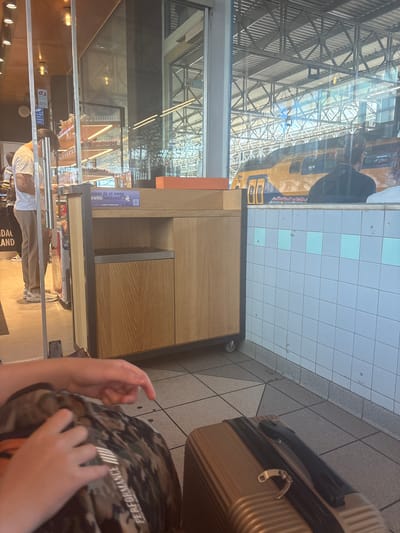 The image captures an indoor scene, likely a waiting area or a shop within a transportation hub, possibly Eindhoven Centraal station. A yellow and blue train is visible through a large glass window, indicating a train station environment. In the foreground, a person's hands are visible, resting on a camouflage-patterned backpack. To the right, a gold suitcase is placed on the tiled floor. An orange object, possibly a box, sits atop a wooden and metal counter unit, which is part of a display or service area. Behind the counter, shelves stocked with various items are visible through a glass partition. A security camera is mounted on a blue sign to the left. The overall lighting suggests an interior space with natural light filtering in from the exterior.
