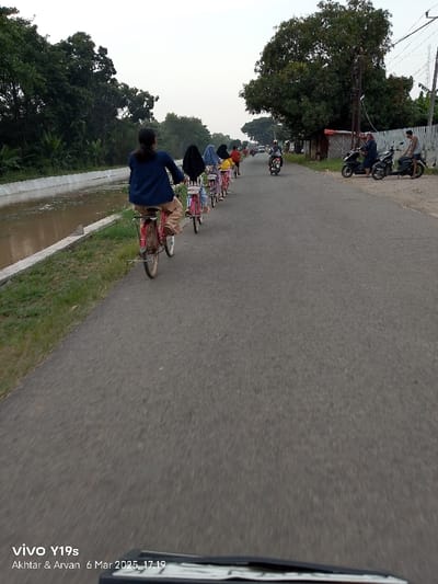 A group of seven people, mostly women, are riding bicycles on a paved road. The road has a canal on the left side. The people are facing away from the camera. The lead cyclist is a woman wearing a blue jacket and beige pants. She is riding a red bicycle with a white seat. The others are dressed in long sleeves and pants. The canal is partially filled with water. The camera is mounted on a moving vehicle. There are some trees on the left side of the road and some more trees and houses on the right. The road appears to be in Kecamatan Gabuswetan, Indonesia.