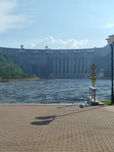This media file captures a wide shot of the Sayano-Shushenskaya Dam in Cheremushki, Russia. The massive concrete dam dominates the background, with several large, circular Russian flags emblazoned on its face. Water cascades down from spillways on the dam, creating a powerful visual of its operational capacity. A wide, blue river flows in the foreground, its surface rippled by currents.
On the right side of the image, a tall lamppost and a signpost with three yellow warning signs are visible. The signs depict symbols of a falling person, a warning triangle, and a power line, indicating potential hazards. A metal railing runs along the edge of the riverbank, separating it from a paved walkway.
The walkway in the foreground is paved with tan interlocking bricks. A shadow of what appears to be two people walking is cast upon the bricks. The scene is illuminated by bright daylight, suggesting it is either morning or afternoon. The sky is mostly clear with scattered white clouds. The presence of lush green trees on the left bank of the river and the impressive scale of the dam suggest a remote, possibly mountainous, or hilly natural environment. The overall impression is one of immense engineering power set against a backdrop of natural beauty.