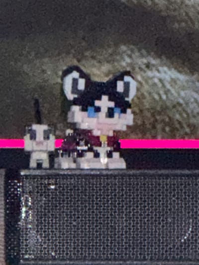 A pixel art representation of a cat, predominantly white with black markings around its ears and a blue eye, is positioned adjacent to a black, textured rectangular object. A vertical pink line bisects the space between the pixel art cat and the rectangular object. The background appears blurry and textured, possibly concrete or a similar rough surface. The location is unknown.