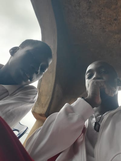 This is a low-angle, close-up selfie taken from a low perspective, showing two young Black men against a backdrop that appears to be an outdoor, possibly covered area with a textured, earthy-toned ceiling or overhang. The lighting is diffused and appears to be daylight, creating soft shadows.
The man on the left is positioned closer to the camera and his face is mostly in shadow, but his mouth is slightly open, revealing teeth, and he has a faint smile. He is wearing a white garment, possibly a hoodie or jacket, and a bit of a red garment is visible in the lower left corner, possibly pants or a shirt. His left ear is visible.
The man on the right is slightly further back and his face is more clearly visible. He is resting his right hand on his chin and looking directly at the camera. He has a neutral expression. He is wearing a white zipped jacket over a white shirt, and a dark pendant necklace is visible on the shirt.
The environment suggests an urban or semi-urban setting in Nsugbe, Nigeria. The rough, textured ceiling could be part of a building's architecture, a market stall, or an outdoor recreational structure. The presence of a blue plastic chair is visible between the two men, suggesting a casual, perhaps public, gathering place. The overall impression is of a candid moment captured by the individuals themselves.