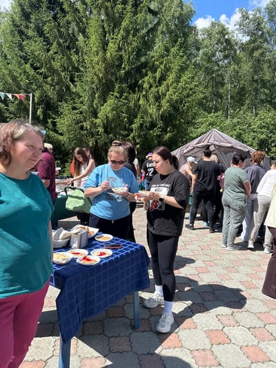 In Duvankul', Russia, a group of people are gathered outdoors, possibly for a community event or market. The scene is set with a backdrop of lush green trees and a clear blue sky with scattered clouds, suggesting a pleasant, sunny day, likely late morning or afternoon. The ground is paved with light-colored interlocking stones.

Several individuals are interacting, with some standing under a tent that appears to be set up for shade. One woman in a black t-shirt is holding what looks like food in a container and a small utensil. Another woman in a blue t-shirt is examining something in her hand, also appearing to be food. A table draped with a dark blue tablecloth displays several plates with slices of pie and small bowls of food, indicating a food service or sampling station. Among the objects visible are white paper plates, napkins, and a smartphone.

The overall mood seems relaxed and casual, with people engaged in conversation and enjoying the outdoor setting. Some individuals are casually dressed, wearing t-shirts and comfortable pants. The presence of a tent and a food table suggests an organized event, perhaps a local festival, fair, or a gathering of some sort. The people's focus on the food and their interactions imply that eating and socializing are the primary activities. There is some visible text on the black t-shirt of the woman in the foreground, but it is not entirely legible in the provided image. The image captures a snapshot of a community enjoying a warm day together.