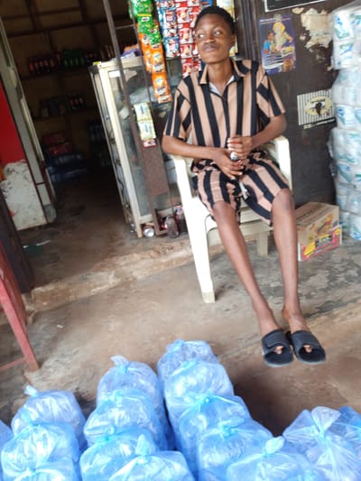 The image depicts a young person sitting outside or at the entrance of a small shop in Umuahia, Nigeria. The individual, dressed in a distinctive black and tan striped short-sleeved shirt and matching shorts, is seated on a white plastic chair. They hold a small, dark object in their hands, possibly a bottle or a phone, and gaze towards the left with a neutral, slightly distant expression. Black slide sandals are visible on their feet.
The setting appears to be a local convenience store or kiosk. To the left of the person, the shop's interior reveals shelves stocked with various beverages in bottles and cans, along with numerous packets of snacks, including brands like "Mamee" and "Chikki," hanging from display racks. A glass display cabinet separates the seating area from the main shop space. The ground is a rustic, light brown surface, likely concrete or packed earth.
To the right of the individual, several large, clear plastic bags filled with smaller, individually packaged sachets of water are stacked against a wall. A cardboard box, bearing the yellow text "SWEET," rests on the ground near the person's feet. More bags of sachet water are arranged across the foreground of the image. A poster with illustrations and some unreadable text is taped to the wall behind the person, next to a black-and-white striped poster featuring the outline of the African continent. The scene is brightly lit, indicating it is daytime. The overall impression is of a casual, everyday commercial setting typical of a neighborhood shop in the region.