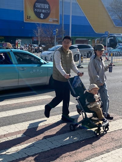 A medium, outdoor, eye-level shot shows a family pushing a stroller in Taihe District, China. The father is pushing the stroller, wearing a white long-sleeve shirt, tan vest, black pants, and black shoes. A second adult, possibly the mother, is following, wearing a grey sweatshirt, blue jeans, and a green baseball cap. Their child is sitting in the stroller wearing a tan hat, brown coat, and brown pants. They are walking across a crosswalk that is worn. To the left of the father is a light blue vehicle. Behind the family, there are rows of parked cars. Behind the cars is a light blue building with a large yellow sign on the side with black lettering. The sign reads "星聚會", "不仅仅是KTV", "快乐专聚会", and "原来……玩期付". There are also English words "KTV" and "1F". There is a white and blue sign behind the mother and child, as well as a streetlight. The sky is light blue, and it is sunny.