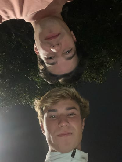 The inverted image captures two young adult males in a close-up, low-angle selfie at night in Aldoar, Foz do Douro e Nevogilde, Portugal. The upper subject (appearing at the top if rotated 180 degrees) has dark brown, medium-length hair and wears a light pink t-shirt. He faces the camera with a subtle smile. Below him (appearing at the bottom if rotated) is a male with light brown, short hair, wearing a light-colored collared jacket with a zipper. He exhibits a subtle smile and a small hoop earring in his visible right ear. The background is dark, consistent with nighttime, and includes blurred dark green foliage.
