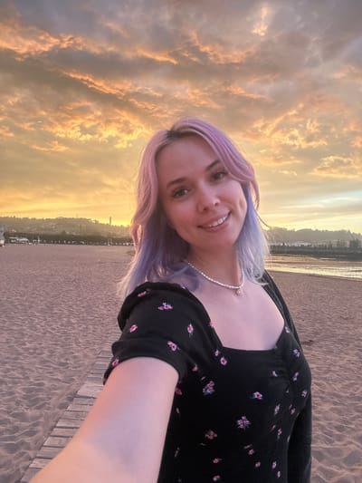 A woman with purple-tinted hair is taking a selfie on a sandy beach. She is wearing a black dress with a floral pattern and a pearl necklace. The beach is wide with wooden planks leading away from the viewer. In the background, there is a calm body of water with a shoreline featuring buildings and hills under a dramatic sky filled with orange and pink clouds, indicative of a sunset. The scene is likely located in Gijón, Spain, possibly near the neighborhood of El Arbeyal, given the expanse of the beach and the view of the coastal city.