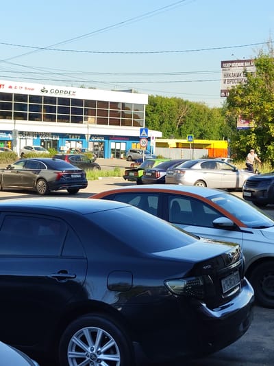 The image shows a street scene in Zhukovsky, Russia, with several cars parked on the side of the road. In the foreground, a black Toyota sedan is parked facing away from the camera. To its right, a silver sedan is visible. Further back, other vehicles, including a dark gray sedan and a green car, are also parked. A billboard with Russian text is visible in the background, along with trees and buildings. The sky is clear and blue, indicating it is daytime. There is a pedestrian crossing and a speed limit sign in the middle ground.