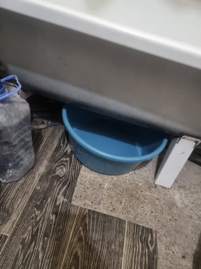 A blue plastic basin is positioned beneath the edge of a white bathtub, collecting what appears to be dripping water. To the left of the basin, a large, clear plastic bottle filled with a dark liquid and a blue lid is visible. The floor beneath these objects is a mixture of dark wood-patterned linoleum and a speckled grey concrete-like material. A white metallic object, possibly a conduit cover or bracket, is located to the right of the basin. The overall scene suggests a potential leak or plumbing issue within a domestic setting. The location of Saran city administration, Kazakhstan, is not discernible from the provided image.