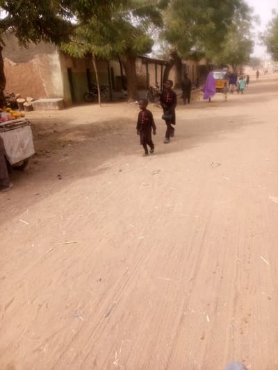 A dusty, sandy road scene features two young boys in matching dark clothing walking away from the camera. Other figures, including one draped in a purple cloth, walk in the same direction. Buildings and trees line the side of the road, and a cart laden with fruit stands to the left. The area suggests a rural or underdeveloped setting. Tire tracks and scattered debris mark the unpaved road.