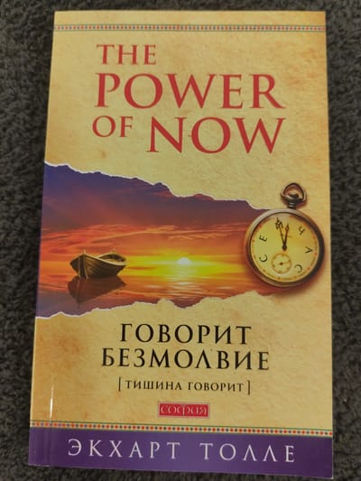 The image is a close-up, overhead shot of a book titled "THE POWER OF NOW" by Eckhart Tolle, lying on a dark grey, textured surface. The book cover features a torn paper effect revealing a sunset scene with a boat on calm water. The title is printed in red lettering. Below the title, there is Russian text that translates to "SPEAKS SILENCE" and in brackets "[SILENCE SPEAKS]". A small red box contains Cyrillic text that reads "СОФИЯ". The author's name, "ЭКХАРТ ТОЛЛЕ", is written in large white letters on a purple band at the bottom of the cover. A vintage pocket watch with Roman numerals is superimposed on the right side of the cover. The book is positioned slightly off-center and appears to be the sole subject of the photograph. There is no indication of the city of Schwäbisch Gmünd, Germany, in the image.