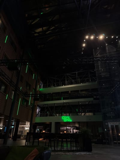 A dark, modern interior space with high ceilings and exposed structural elements. Geometric shapes create a complex web, illuminated by rows of lights. A green neon sign reads "CRAY BAR." Below, a grid-like floor is patterned with illuminated rectangles. In the foreground, a partially visible piece of furniture adds depth to the scene. The overall impression is one of contemporary design and dynamic perspective.